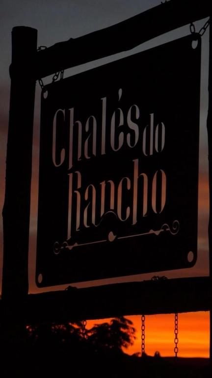 Placa de metal com o nome Chalés do Rancho recortada contra o céu alaranjado do entardecer